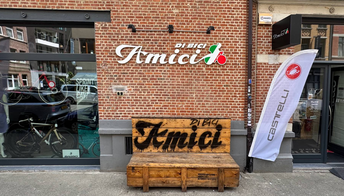 Exterior of a brick bicycle shop named “Amici Di Bici,” with a large logo on the wall, a wooden bench with “Amici” painted on it. A shop window displays bicycles and cycling gear.