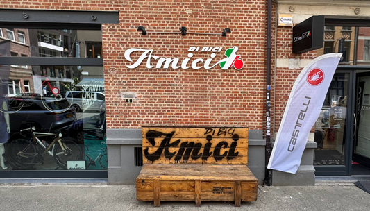 Exterior of a brick bicycle shop named “Amici Di Bici,” with a large logo on the wall, a wooden bench with “Amici” painted on it. A shop window displays bicycles and cycling gear.