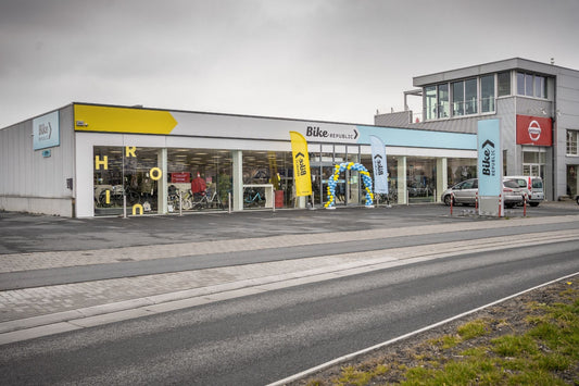 Front view of a modern Bike Republic store with large glass windows, promotional flags at the entrance, and a blue-and-yellow balloon arch on an overcast day.