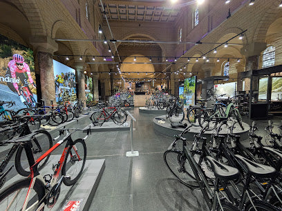 Large bicycle shop with many racing bikes and e-bikes, high arched ceilings, and posters featuring cyclists.